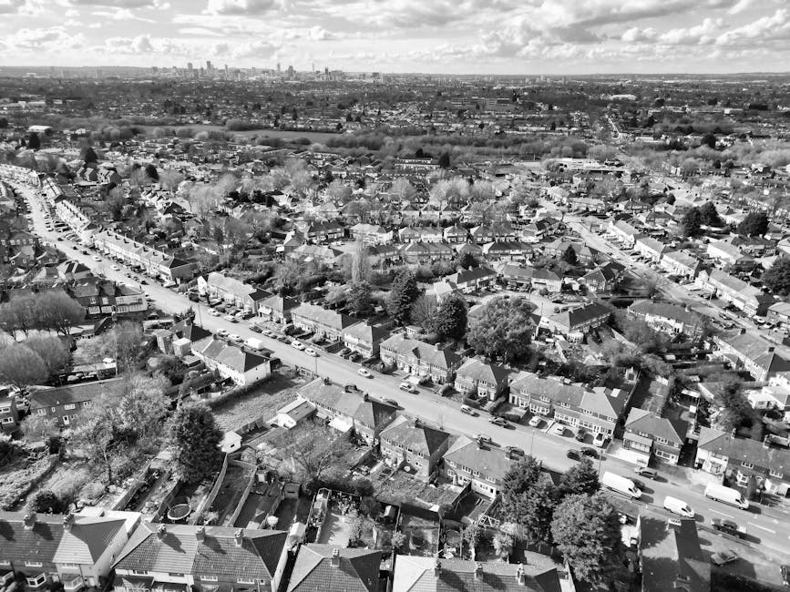 An aerial view of a suburban residential area featuring rows of terraced houses and semi-detached properties with tiled roofs, situated along winding streets and surrounded by small gardens and backya