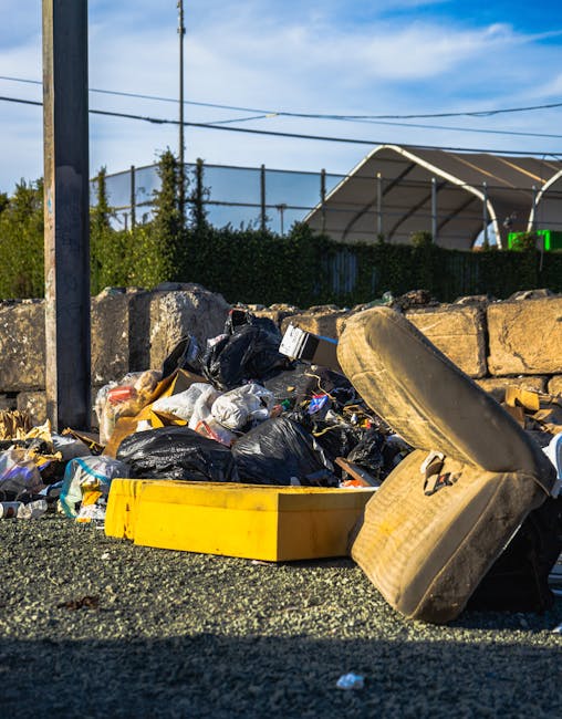 An outdoor scene showing scattered flattened cardboard boxes, primarily green with white star logos, alongside crumpled paper and miscellaneous refuse on a patch of uneven grass and soil. The cardboar