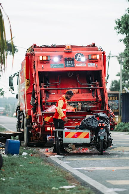 A waste collection worker wearing a high-visibility yellow and red vest is seen operating at the rear of a large red refuse collection truck, which is parked on the side of a paved street. The truck f