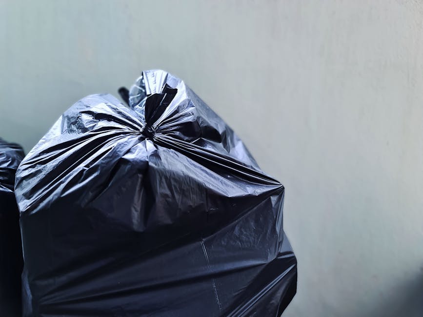 A woman standing on a residential street sidewalk next to multiple black wheelie bins, which are lined up against the brick exterior of a row of terraced houses. The bins have white numbering and are 